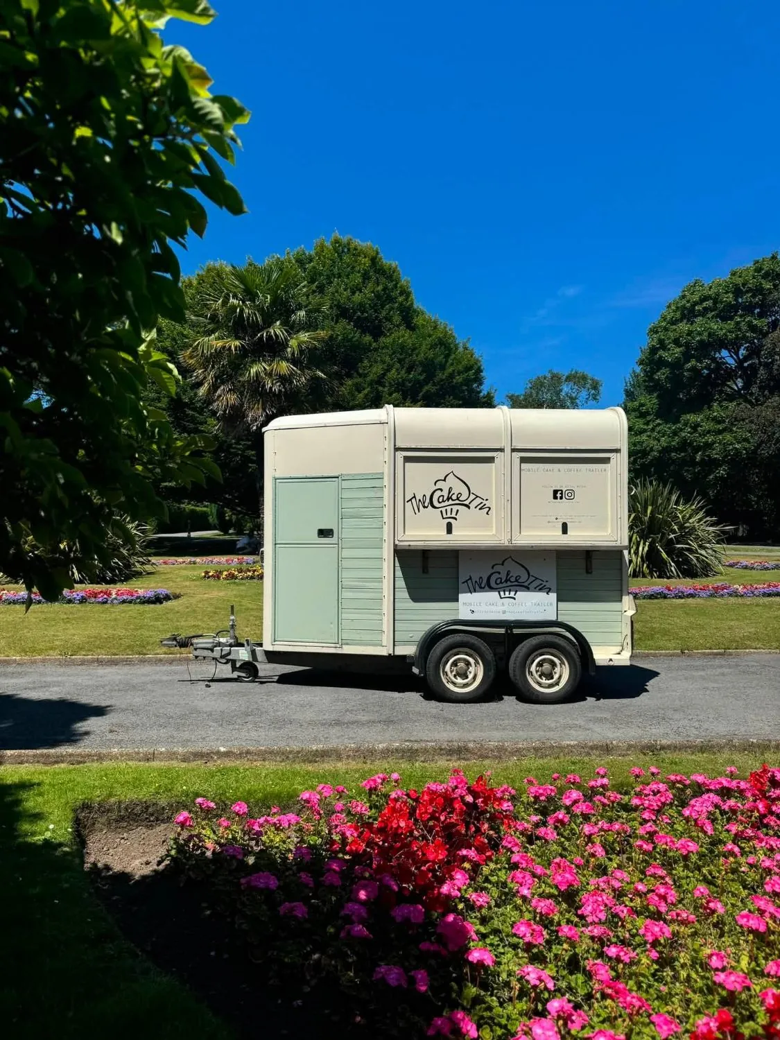 The sage and cream Cake Tin trailer parked beside flowerbeds on a sunny day
